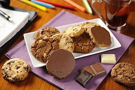 Assiette de cookies au chocolat variés sur une serviette mauve avec carrés de chocolat et tasse de thé sur un bureau.
