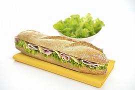 Sandwich baguette garni de jambon, emmental et salade sur serviette jaune, fond blanc lumineux, bol de salade verte.