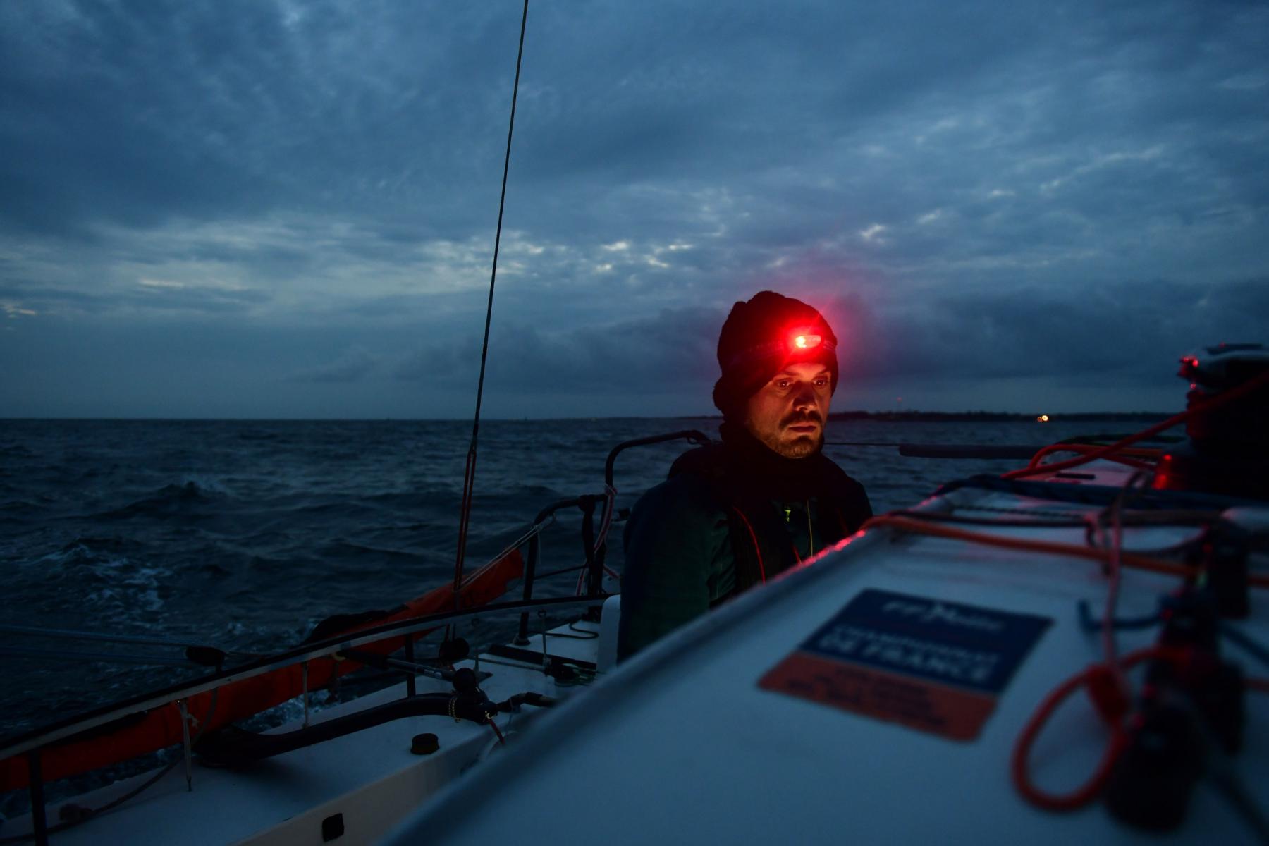 Skipper portant une lampe frontale rouge à bord d’un voilier Mini 6.50 en mer agitée au crépuscule en Bretagne.
