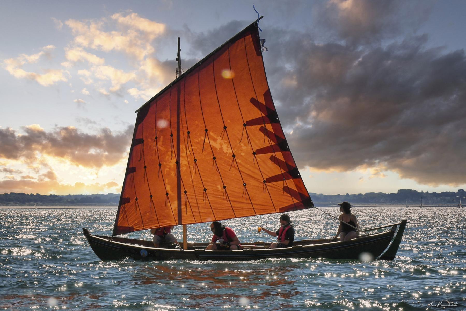 Canot en bois à voile orangée naviguant en Bretagne au coucher du soleil avec quatre passagers à bord.