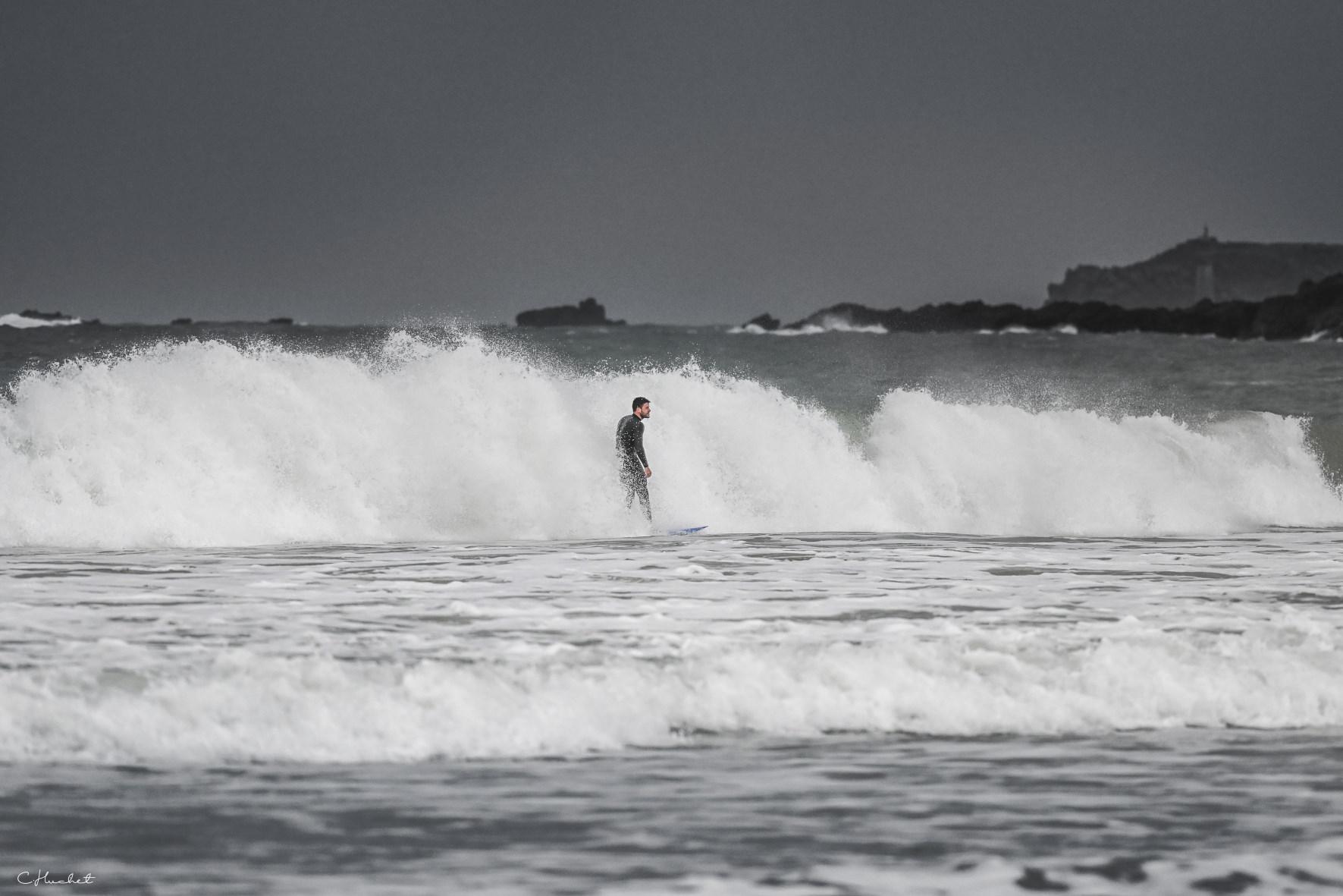 Surfeur en combinaison noire glissant sur une déferlante d’écume blanche sous un ciel gris à la plage de Longchamps.