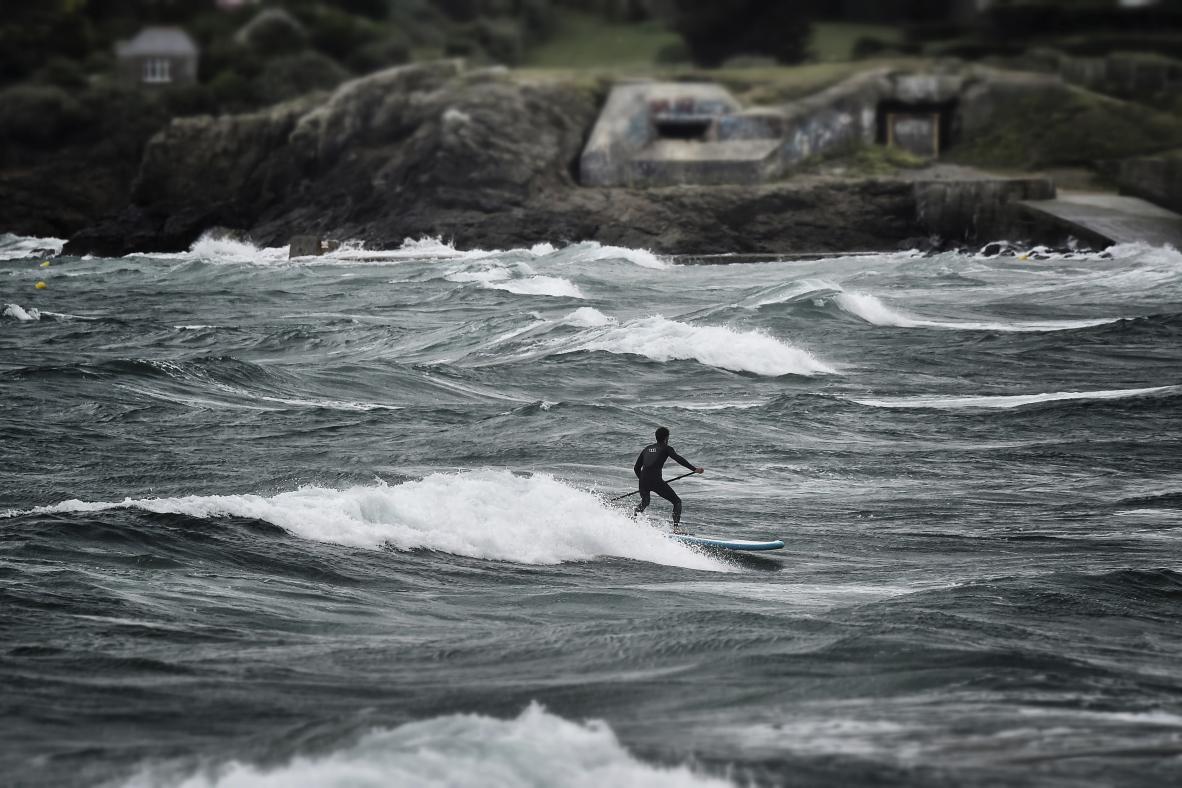 Surfeur en paddle sur une vague écumeuse à Saint-Lunaire devant des rochers et un bunker en arrière-plan.