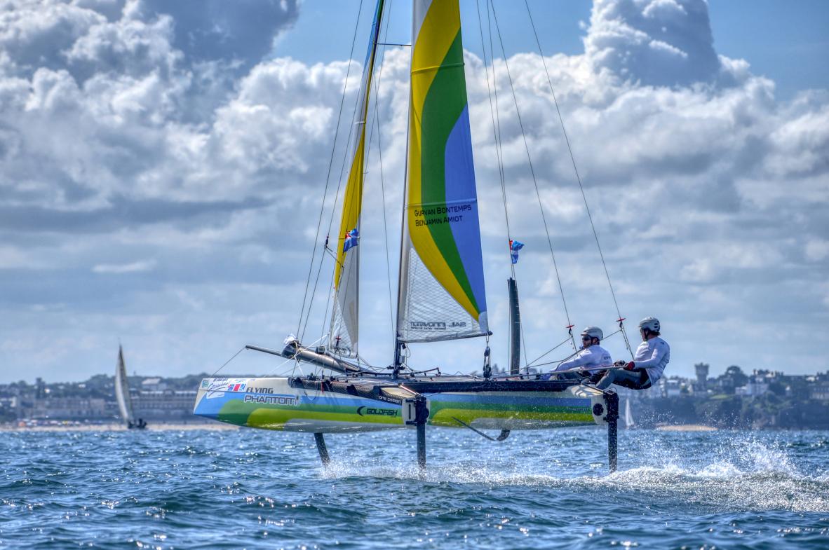 Catamaran Flying Phantom sur foils s’élevant au-dessus de l’eau devant la côte de Dinard sous un ciel nuageux.