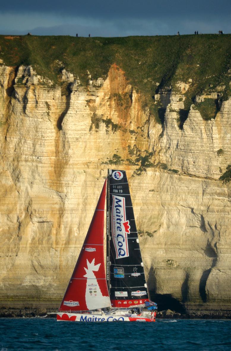 Voilier Imoca Maître Coq aux voiles rouges et noires devant les falaises calcaires du Havre par mer calme.