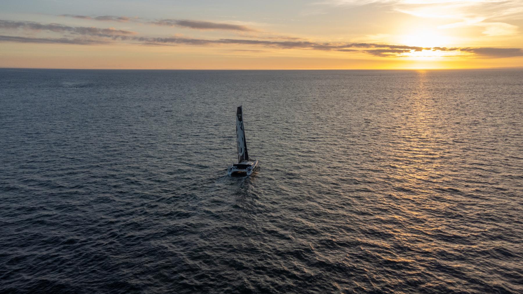 Vue aérienne du catamaran GDD naviguant sur l’océan au coucher du soleil, reflets dorés sur l’eau et horizon dégagé.