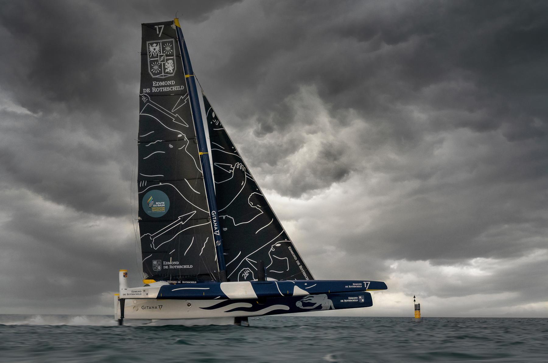 Trimaran Ultim Gitana 17 fendant les vagues sous un ciel d’orage menaçant, voiles sombres et coque bleue.
