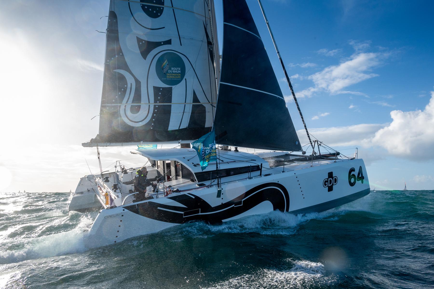Catamaran blanc GDD fendant les vagues au large de Saint-Malo, voiles déployées sous un ciel marin lumineux.