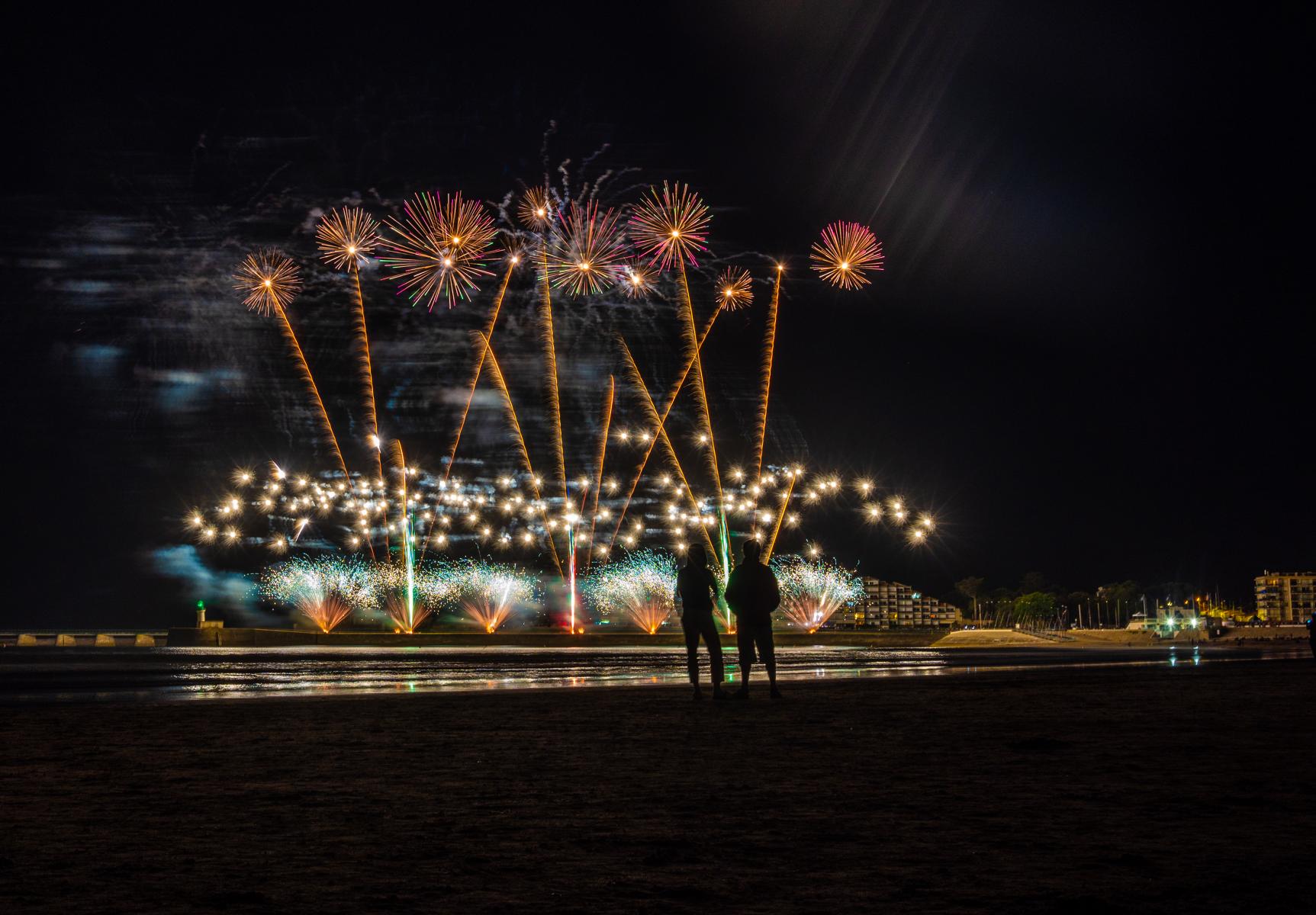 Feu d’artifice doré éclatant au-dessus de l’océan avec silhouettes de spectateurs sur la plage de nuit.