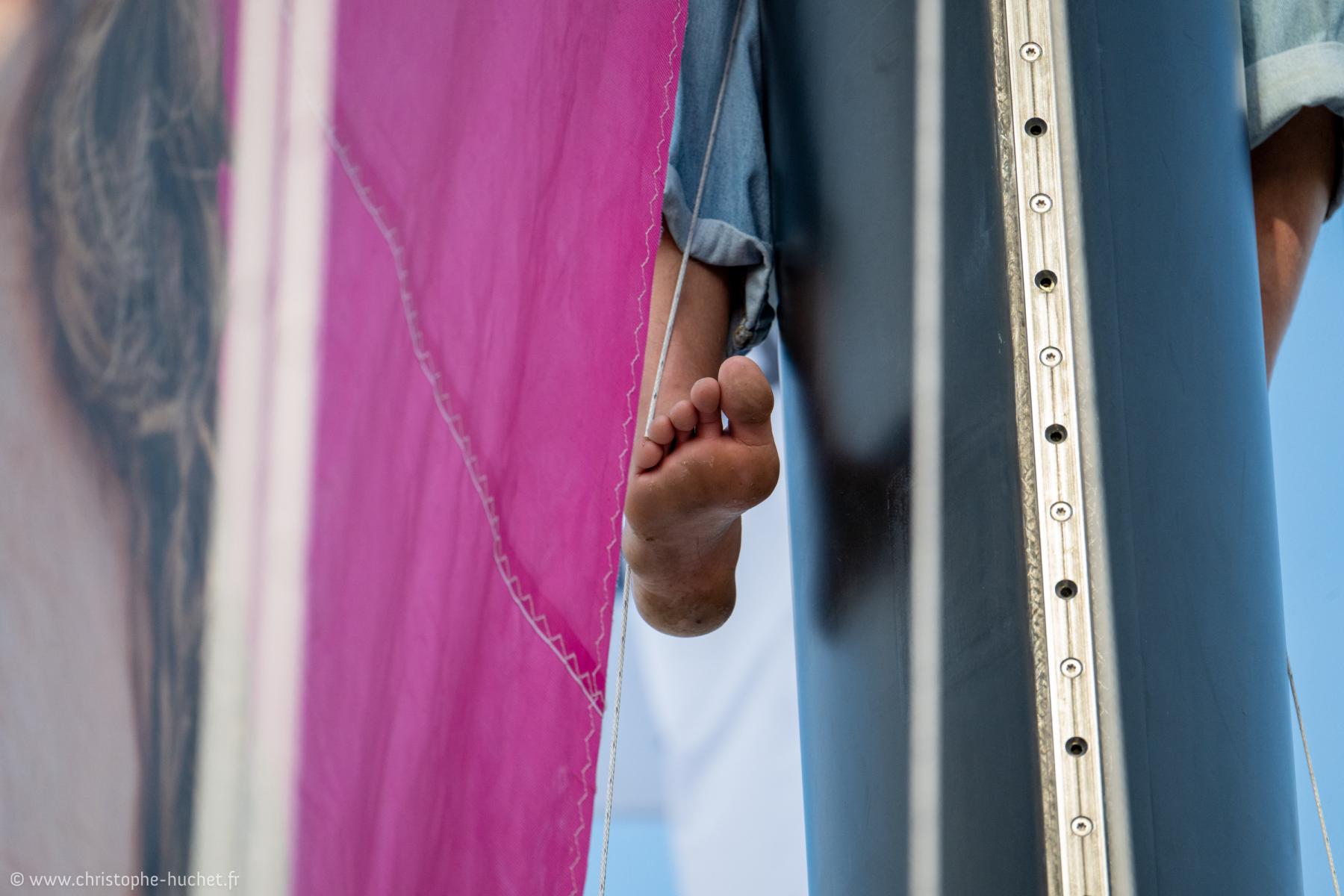 Gros plan d’un pied nu sur le pont de l’IMOCA Lazare, voile rose et mât noir sous un ciel bleu clair.