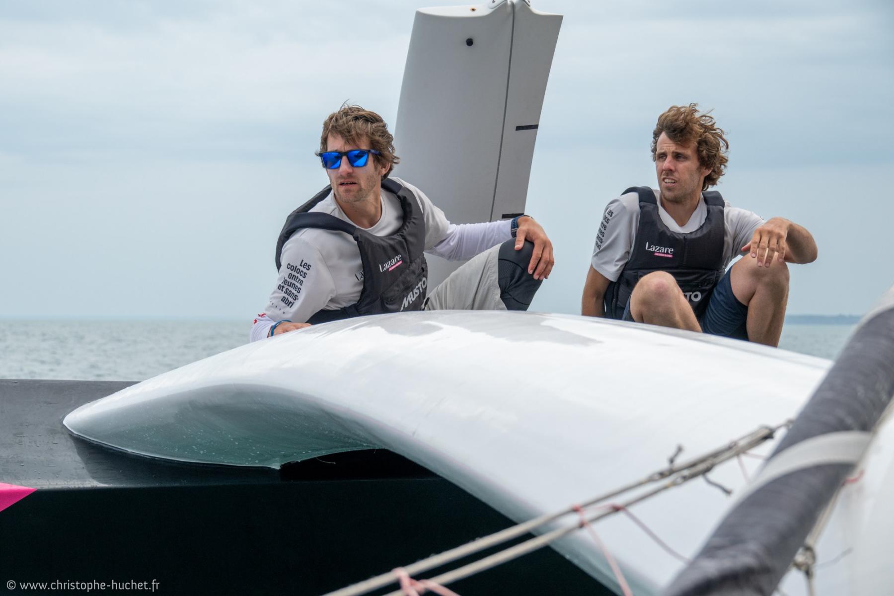 Deux équipiers en gilets de sauvetage sur le pont d’un trimaran blanc Ocean Fifty en mer par temps couvert.