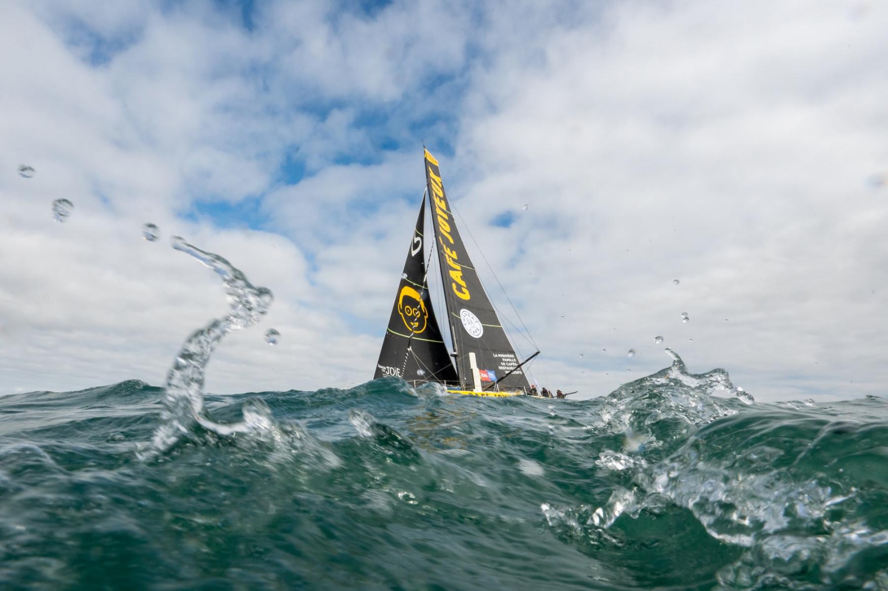 Voilier IMOCA noir et jaune Café Joyeux fendant les vagues à Lorient, éclaboussures d’eau vive au premier plan.