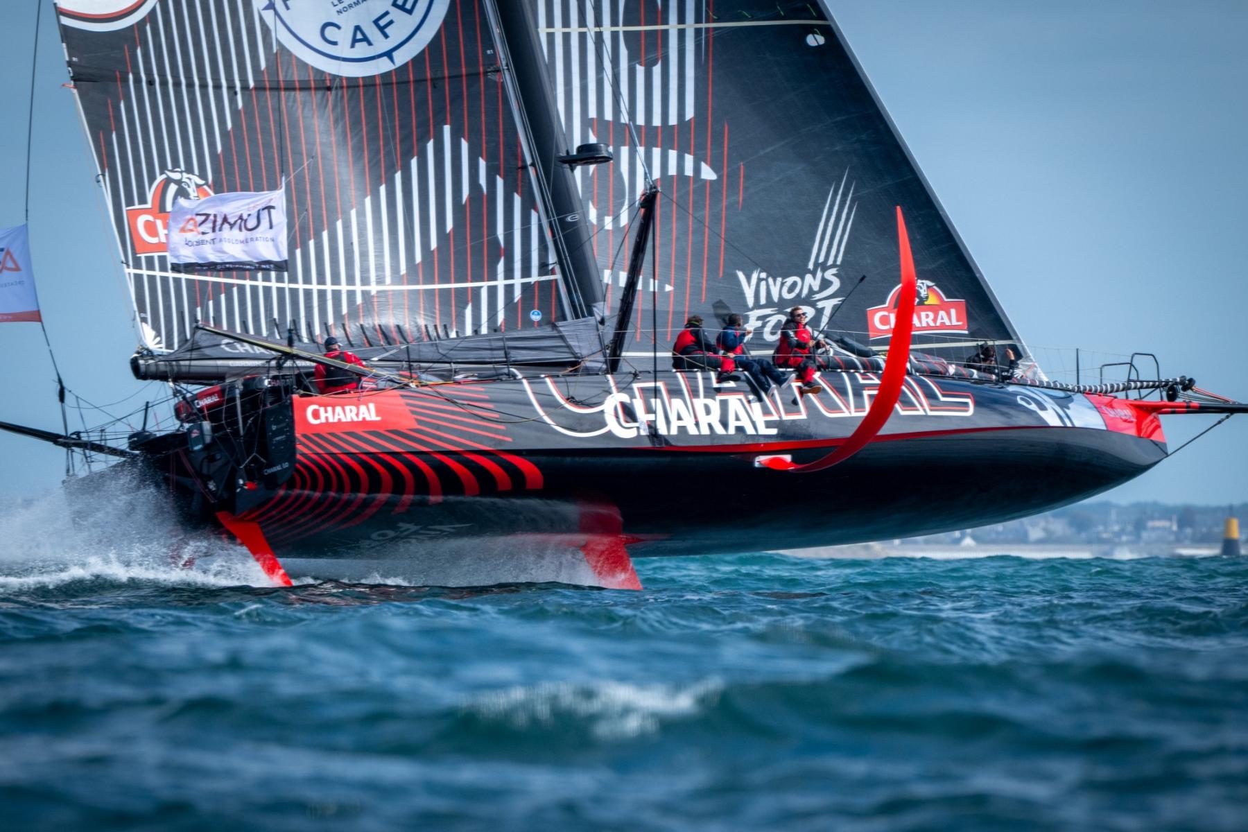 Voilier IMOCA Charal noir et rouge volant sur ses foils au-dessus de l’eau bleue écumeuse lors d’une régate à Lorient.