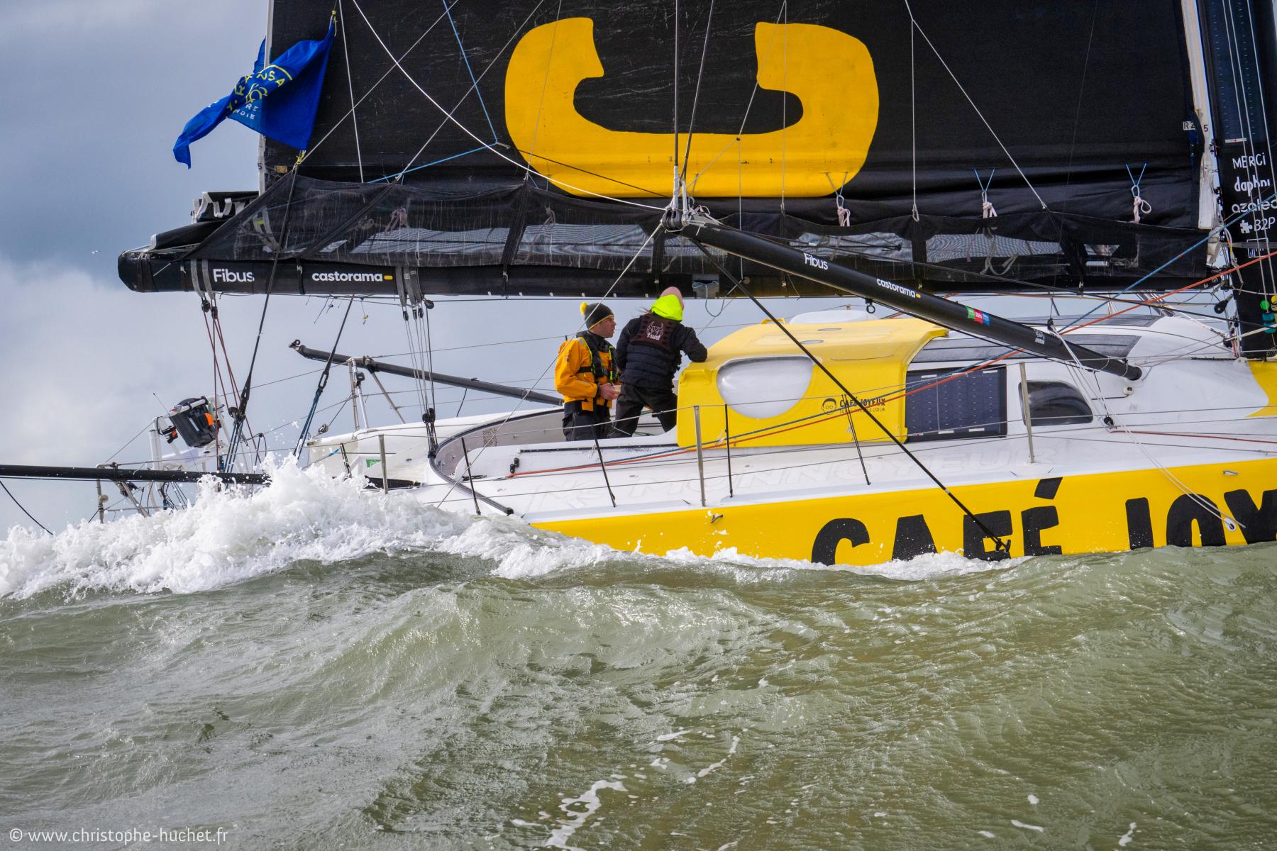 IMOCA Café Joyeux jaune et blanc fendant les vagues au Havre, voile noire avec logo jaune, équipage en action.