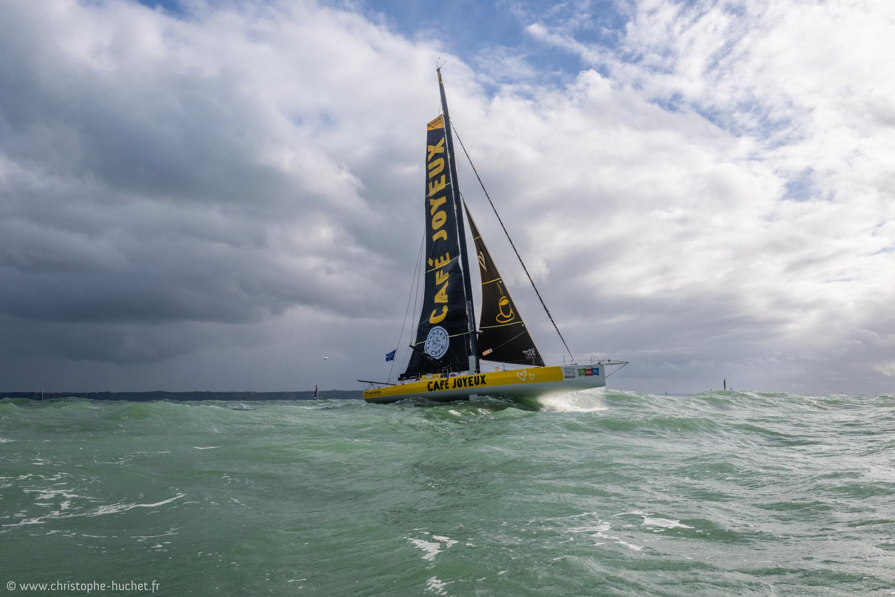 Voilier IMOCA jaune et noir Café Joyeux fendant les vagues vertes sous un ciel nuageux au large du Havre