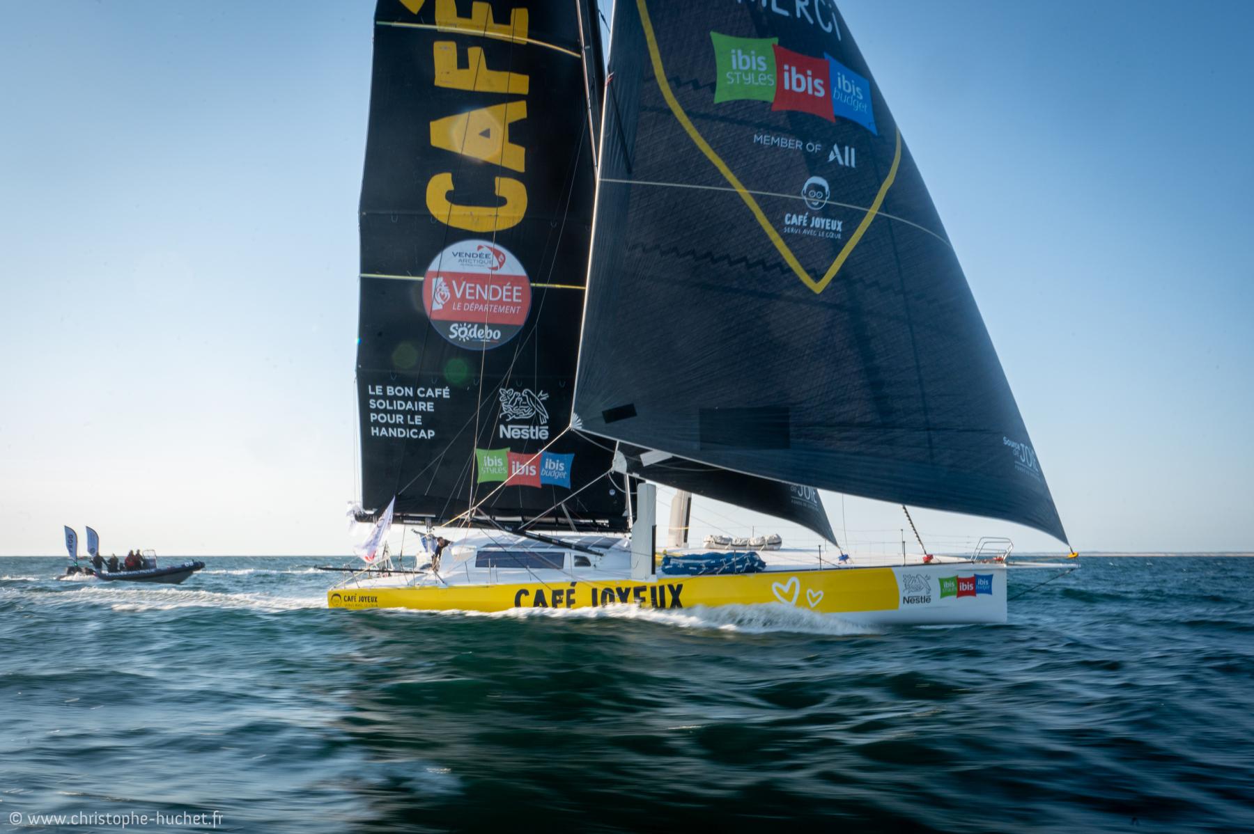 Imoca Café Joyeux aux voiles sombres glissant sur l’eau bleue, ciel clair et horizon dégagé, logos sponsors visibles.