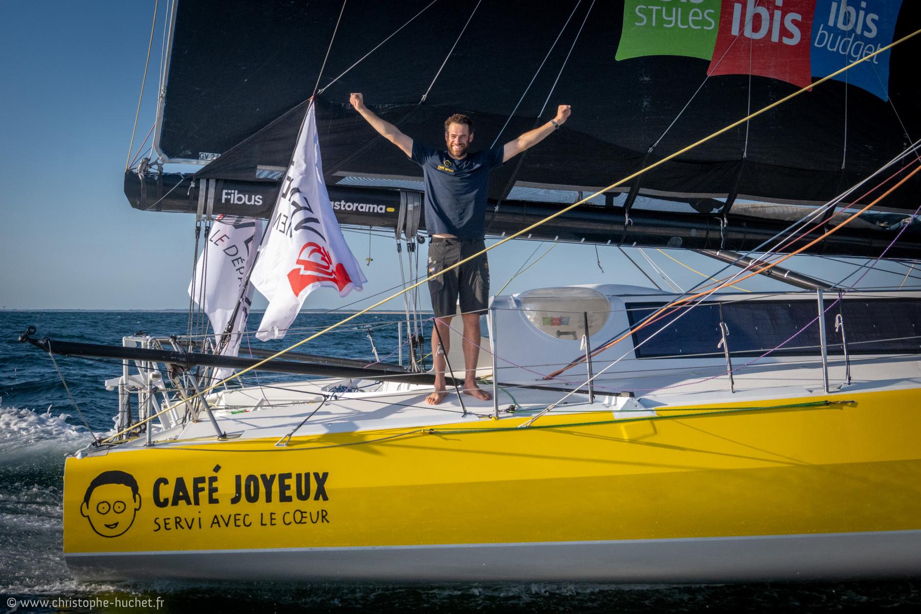 Marin bras levés sur le pont du voilier jaune Café Joyeux, mer bleue et drapeaux de course