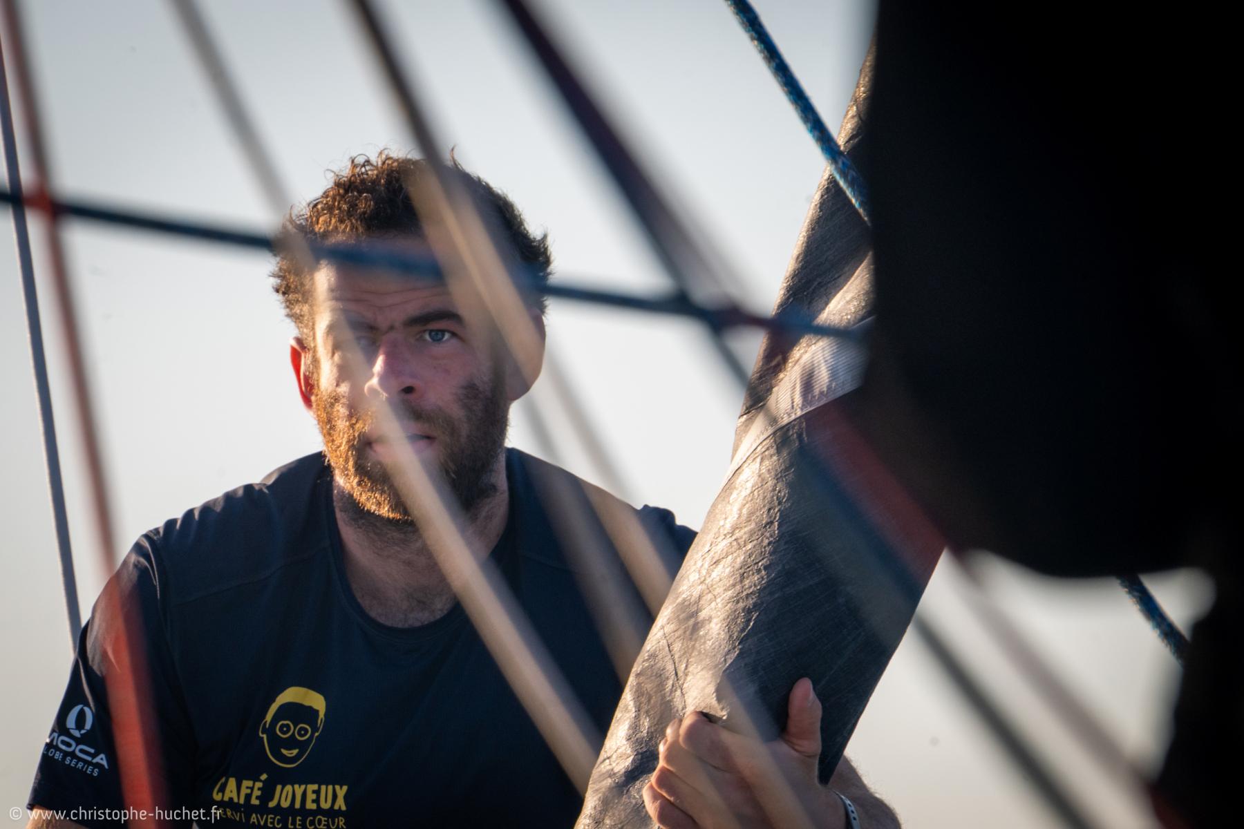 Marin barbu concentré derrière les cordages d’un voilier de course lors d’une arrivée en mer sous un soleil rasant