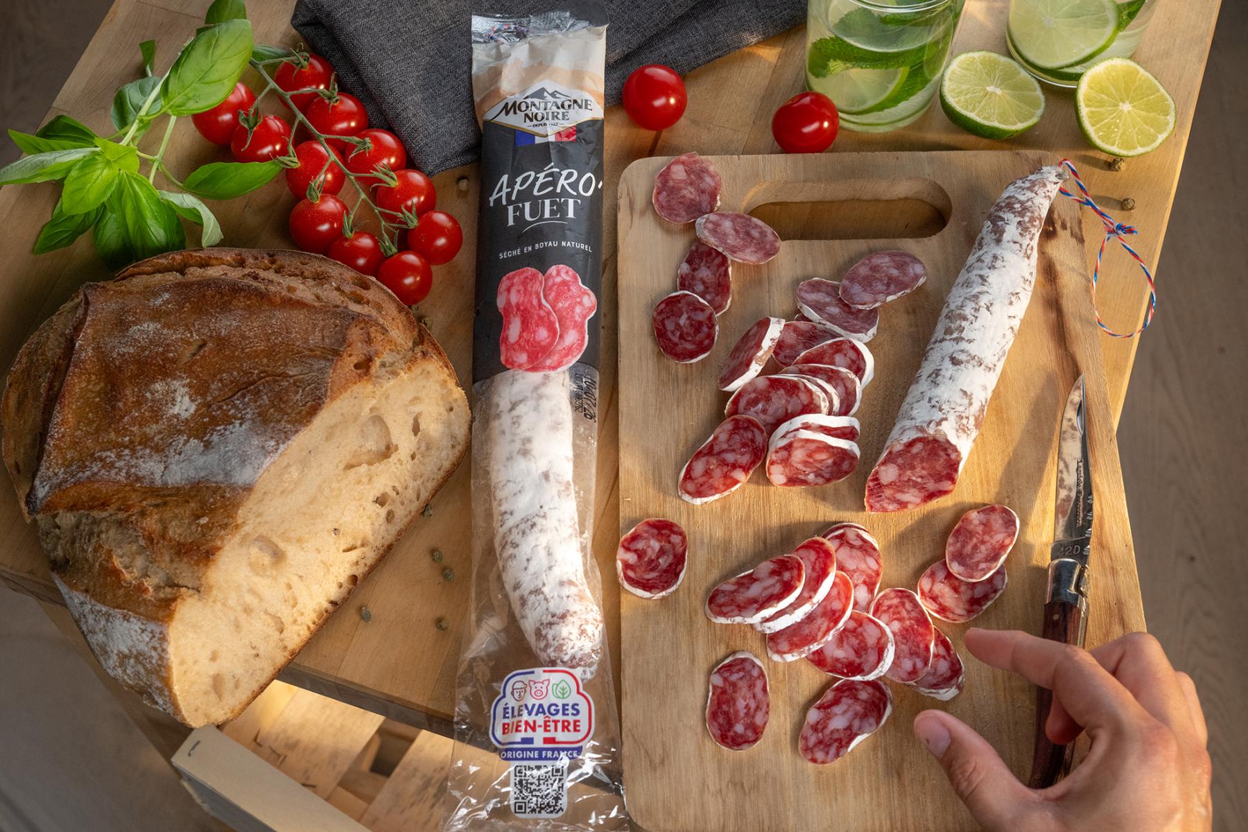 Saucisson fuet tranché sur planche en bois avec pain de campagne, tomates cerises et main sous une lumière chaude